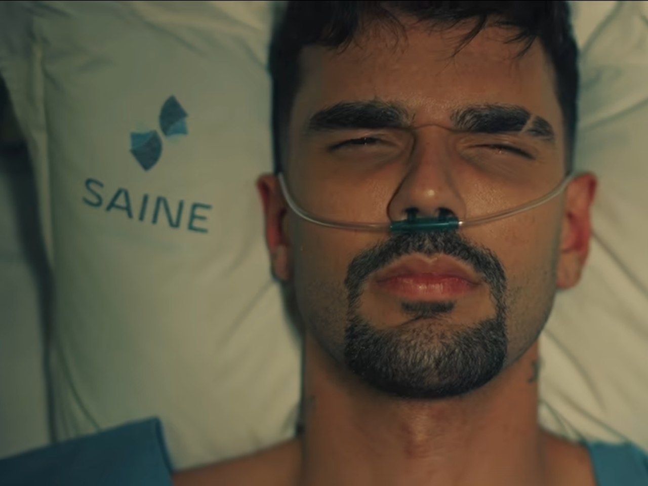 Cena do clipe gravado no Saine, com o cantor deitado em um leito e o travesseiro com o logo do hospital